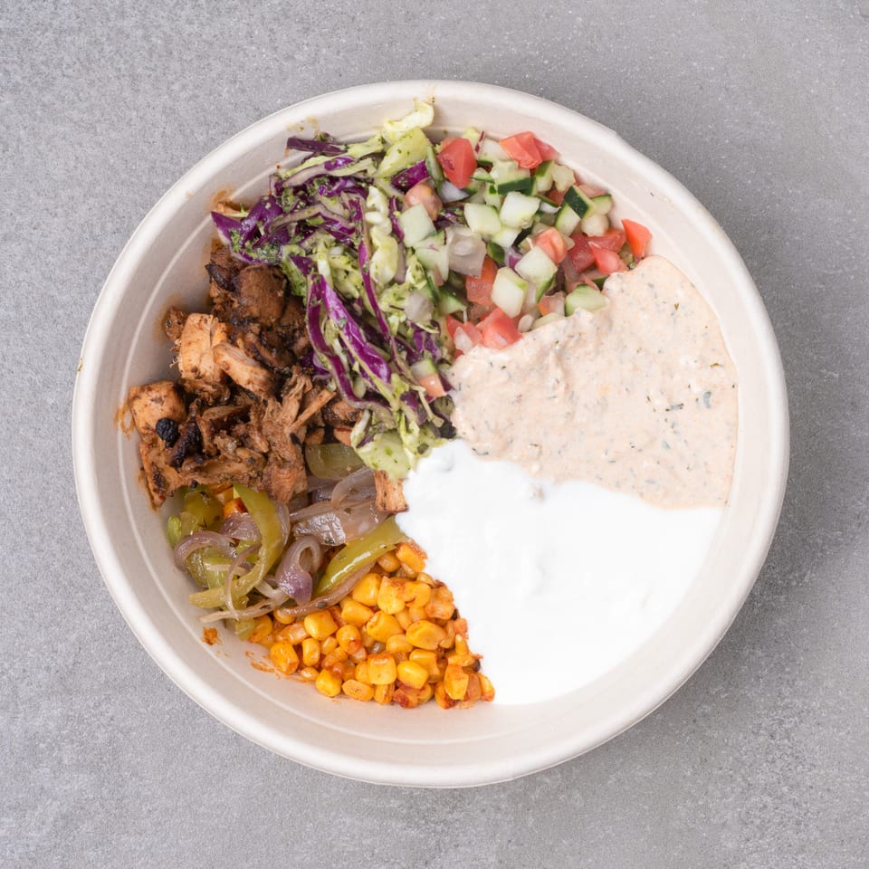 Top-down view of a bowl with grilled chicken, corn, peppers, cabbage, cucumber, tomato, and two sauces.