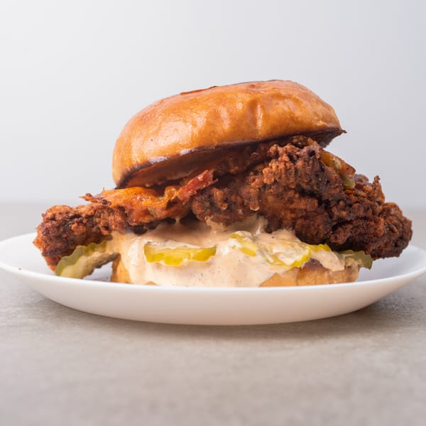 Crispy fried chicken sandwich with pickles and creamy sauce on a brioche bun, served on a white plate.